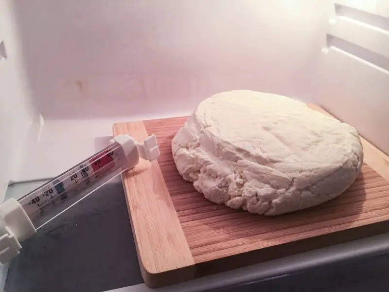 The Parmesan Cheese on a bamboo cutting board inside a refrigerator with a refrigerator thermometer laying off to one side.