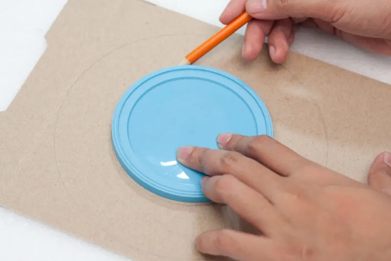 A hand using a pencil to trace around a plastic lid laying on a piece of cardboard.