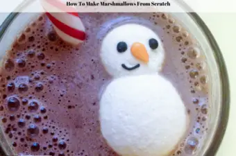 A snowman marshmallow floating in a cup of hot chocolate with a peppermint stir stick beside it.