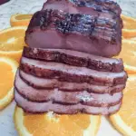 A pre-sliced Frick's Gourmet Ham With Natural Juices glazed with an orange honey sauce and sitting on a platter of orange slices.