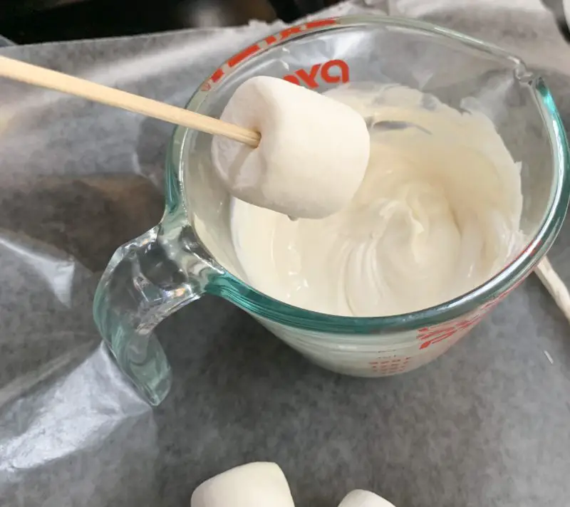 A wooden skewer inserted in a marshmallow about to be dipped in white chocolate.