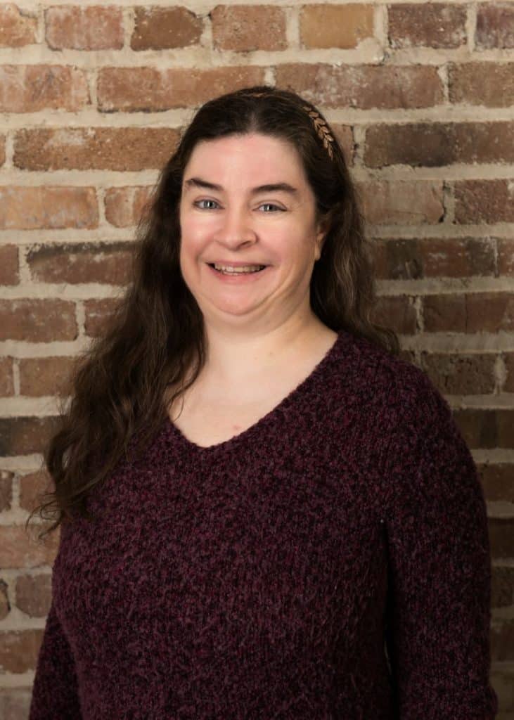 Bright woman smiling in front of brick wall, dressed casually.