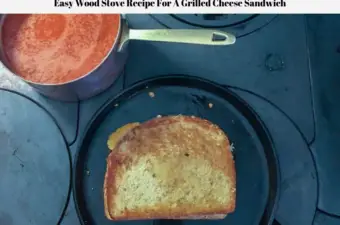 A grilled cheese sandwich cooking in a cast iron griddle on top of a wood burning stove with a pan of tomato soup on the side.