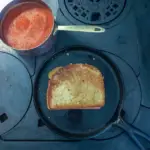 A grilled cheese sandwich cooking in a cast iron griddle on top of a wood burning stove with a pan of tomato soup on the side.